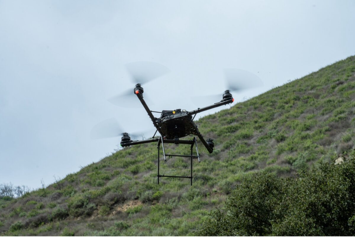 Black quadcopter drone with spinning propellers hovers in flight against a clear sky, with a green hillside in the background. The drone features a frame attachment beneath its body for carrying equipment or payloads.